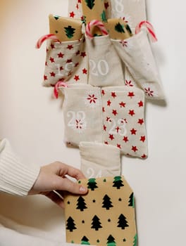 The hand of one Caucasian unrecognizable teenage girl takes a gift lying next to an advent calendar near a pocket with the number 24 in the evening in the living room, close-up side view.
