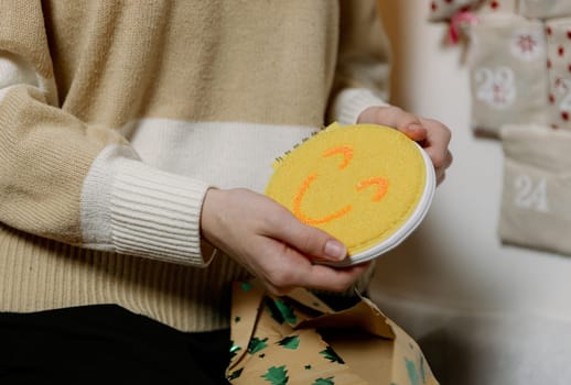 One Caucasian unrecognizable teenage girl holds in her hands an unwrapped gift yellow notepad with a smile from an advent calendar hanging on a white wall in the evening in the living room, close-up side view.