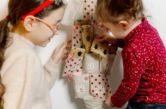 Two beautiful and happy Caucasian girls happily take out surprise gifts with their hands from the pockets of a wall advent calendar hanging on the wall near the Christmas tree early in the morning in the living room, close-up side view.