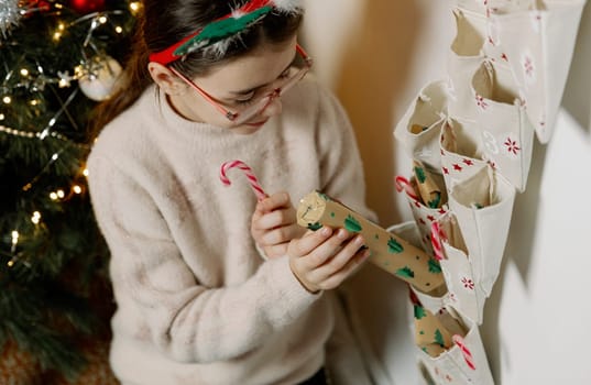 One Caucasian girl with a headband of deer antlers takes out with her hands a surprise gift and a lollipop candy from the pocket of a wall advent calendar hanging on the wall near the Christmas tree in the evening in the living room, close-up side view from above.