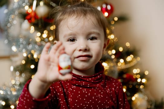 One beautiful Caucasian baby girl shows a gift, taking it out of the pocket of a wall advent calendar, standing by a blurry Christmas tree in the evening in the living room, close-up side view.