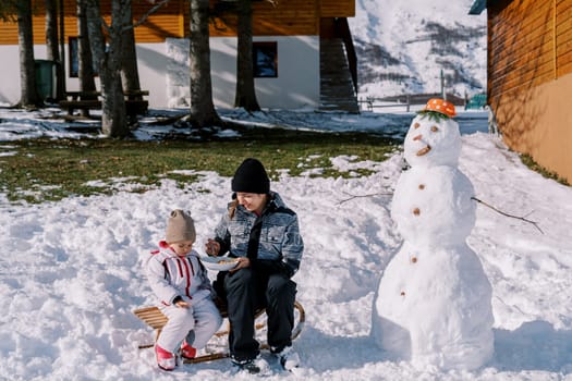 Mom feeds a little girl from a spoon, sitting on a sleigh near a snowman near a cottage. High quality photo