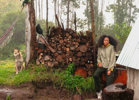 young tourist with afro hair sitting at dawn next to a big block of firewood and a white dog in a natural and rural environment. tourism day. High quality photo