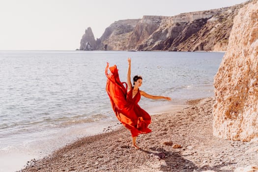 Woman red dress sea. Female dancer in a long red dress posing on a beach with rocks on sunny day. Girl on the nature on blue sky background