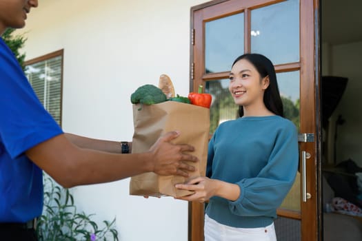 Asian deliver man worker handling bag of food, fruit, vegetable give to young beautiful female in front of the house.