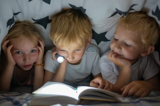 Three young children are reading a book with a flashlight under the covers at night