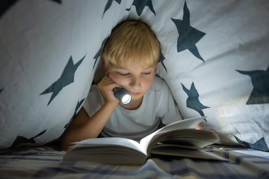 A little boy reads a book with a flashlight under the covers at night
