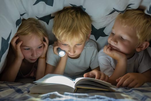 Three young children are reading a book with a flashlight under the covers at night