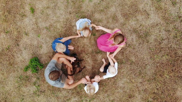 A friendly family sit holding hands on the grass with a dog. View from the drone
