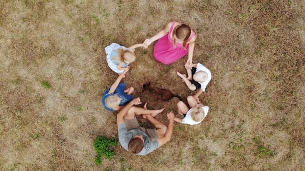 A friendly family sit holding hands on the grass with a dog. View from the drone