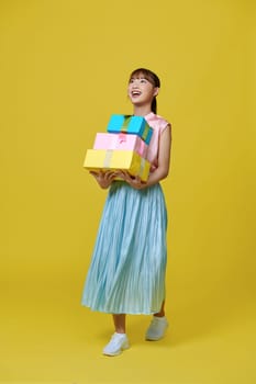 Attractive young girl in dress holding stack of gift boxes isolated over yellow background