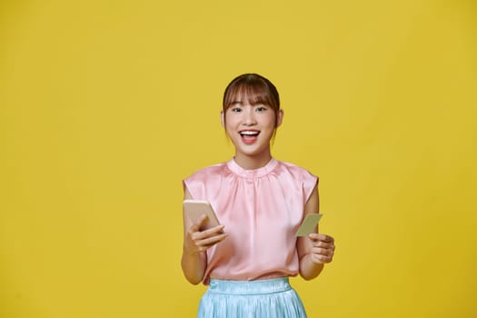 Cheerful young girl showing plastic credit card while using mobile phone 