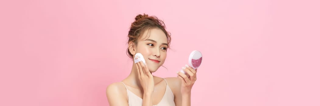 Beautiful young woman applying face powder with puff applicator on pink background