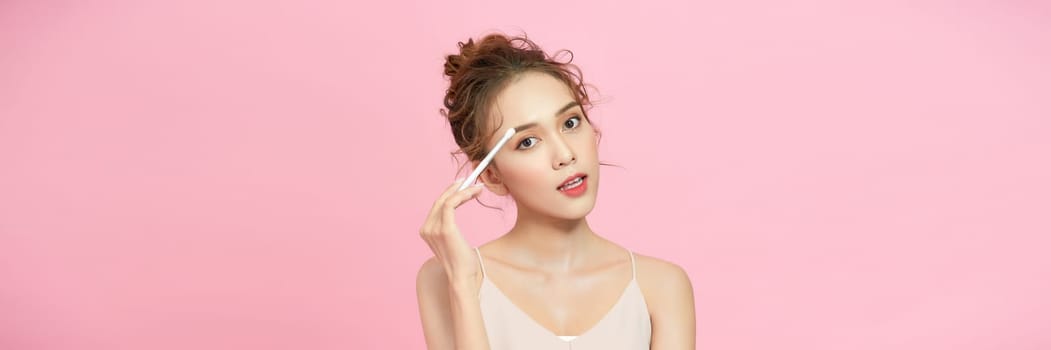 Woman applying face powder on nose making makeup using cosmetic brush posing over pink