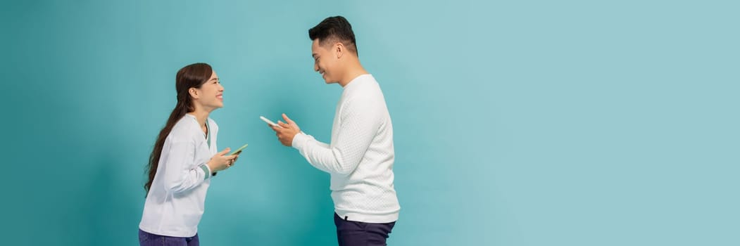 Asian girl and guy using phones sending and reading messages, standing against blue background facing each other