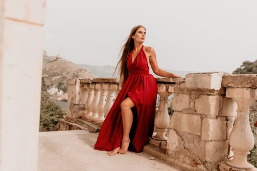 Side view a Young beautiful sensual woman in a red long dress posing on a volcanic rock high above the sea during sunset. Girl on the nature on overcast sky background. Fashion photo