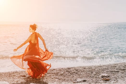 Side view a Young beautiful sensual woman in a red long dress posing on a rock high above the sea during sunrise. Girl on the nature on blue sky background. Fashion photo.