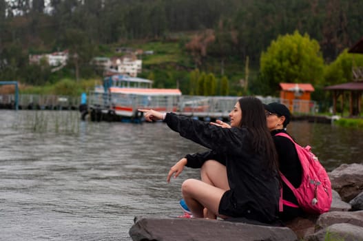 two friends sitting on a wet rock on the shore of a lagoon full of water and one of them pointing to the horizon. backpacker's day. High quality photo