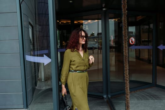 Woman leaves a supermarket. Caucasian model with long brunette hair, wears sunglasses and a khaki dress.