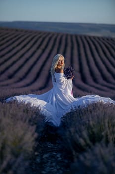 Blonde woman poses in lavender field at sunset. Happy woman in white dress holds lavender bouquet. Aromatherapy concept, lavender oil, photo session in lavender.