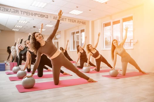 A group of six athletic women doing pilates or yoga on pink mats in front of a window in a beige loft studio interior. Teamwork, good mood and healthy lifestyle concept