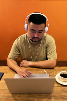 Vertical image of Asian man with headphones using laptop working in coffee shop. Orange wall background. Freelance and digital nomad concept.
