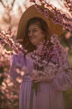 Woman blooming peach orchard. Against the backdrop of a picturesque peach orchard, a woman in a long pink dress and hat enjoys a peaceful walk in the park, surrounded by the beauty of nature