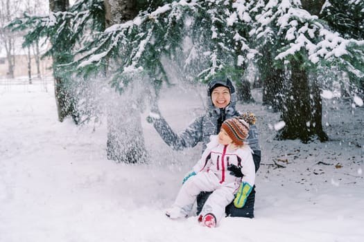 Smiling mom with a little girl sits on her knees in the snow under snowfall from pine branches. High quality photo