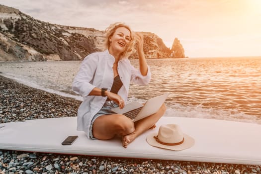 Successful business woman in yellow hat working on laptop by the sea. Pretty lady typing on computer at summer day outdoors. Freelance, travel and holidays concept.