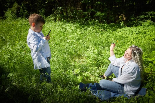 A young teenage son takes pictures of his mother on a cell phone in nature. Mom and boy having fun in the grass in clearing