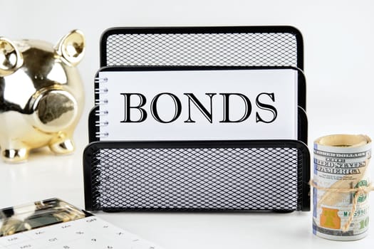 BONDS text on a white piece of paper next to a piggy bank, a roll of money and a calculator on a white background