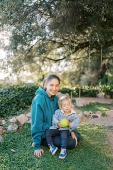 Smiling mother with a little girl with a ball in her hands on her knees sits on green grass. High quality photo