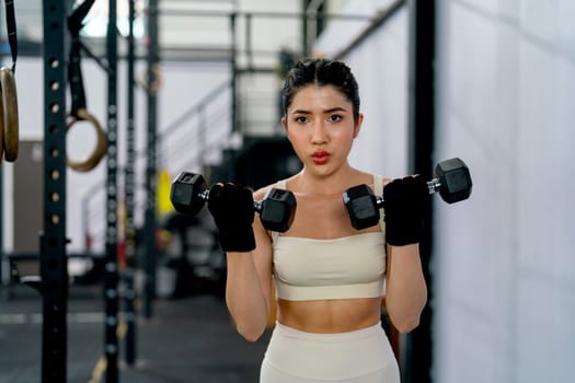 Pretty Asian sport woman hold and lift two dumbbells up and look forward in fitness gym and look concentrate to training.