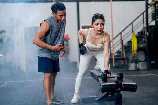 Wide shot of Asian sport woman sit and stay in position of exercise with hold dumbbells and look forward also support with trainer or coach in fitness gym.