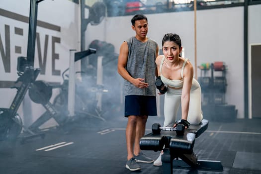 Wide shot of Asian sport woman sit and stay in position of exercise with hold dumbbells and look forward also support with trainer or coach in fitness gym.
