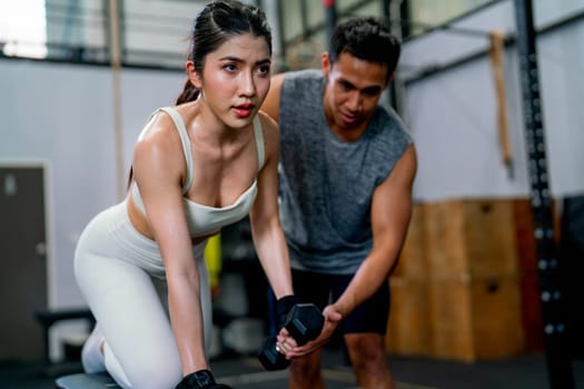 Asian sport woman sit and stay in position of exercise with hold dumbbells and look forward also support with trainer or coach in fitness gym.