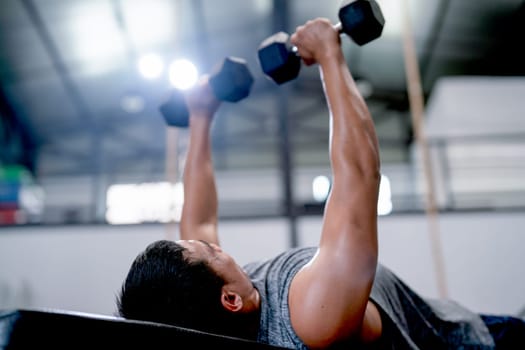 Close up Asian sport man lift dumbells up to over head during lie on long chair in fitness gym.