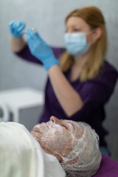 Blurred background. In the background you can see the blurry figure of a nurse scooping a preparation from a vial into a syringe. In the foreground lies a woman with her face wrapped in foil. The woman is being prepared for a procedure.