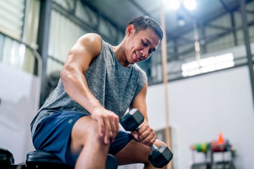 Asian sport man sit and hold dumbbell on chair in fitness gym and look relax and smiling after train the body for healthy lifestyle.