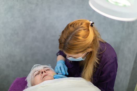 The patient is lying on a bed in an aesthetic clinic, with a lamp lighting over her. The woman has a grimace of pain on her face as she has needle mesotherapy performed.