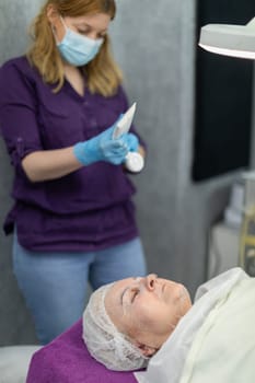 A beautician stands by a cosmetic bed and squeezes cream from a tube. Next to her lies a woman who has her eyes closed. She is wearing a sanitary cap on her head.