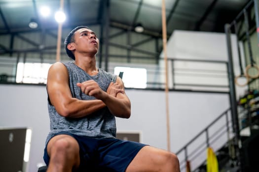 Asian sport man with grey vest sit on chair and action of relax or stretching arm in fitness gym.