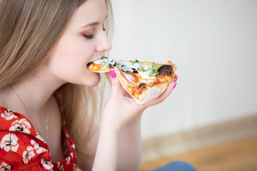 A young girl holds a slice of pizza. She bites into the pizza with pleasure. The woman's eyes are closed. She is sitting on the floor, and a white wall can be seen in the background.