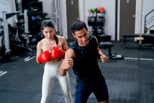 Wide shot of Asian sport trainer man help woman to practice with boxing to punch and hit for good action and movement in fitness gym.