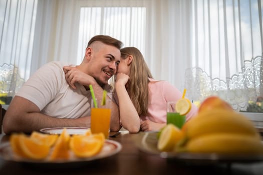 In the foreground, blurred fruit plates standing on a table. Behind the table sits a boy with a girl. The girl whispers something in the ear of the boy. In the background you can see a window covered with a white curtain.