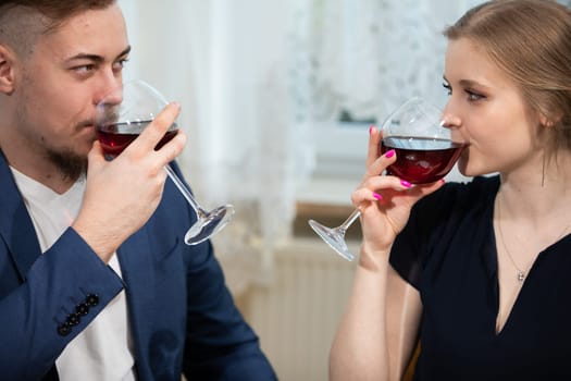 A man and a woman are sitting against a window. The window is obscured by a curtain. The couple is holding glasses and drinking red wine. They are dressed elegantly.