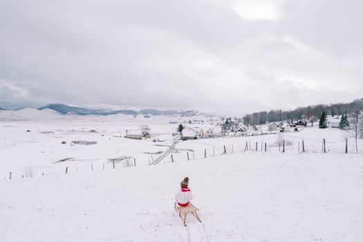 Little girl rides a wooden sled down a hill on the outskirts of a village. Back view. High quality photo
