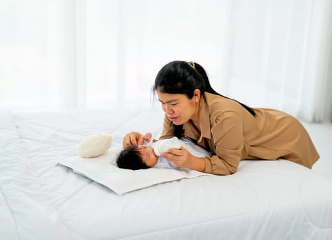 Young Asian mother give milk in bottle to newborn baby lie on bed and take care with happiness in bedroom of their house.