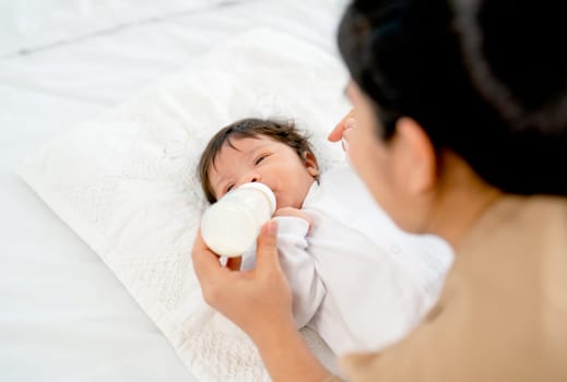 Back of Young Asian mother give milk in bottle to newborn baby lie on bed and take care with happiness in bedroom of their house.