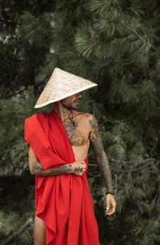 Sexy male monk in a red cape and an Asian triangular hat with tattoos in nature